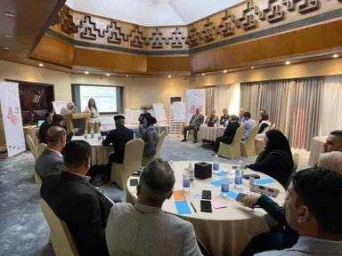 A conference room with people seated at round tables and two women presenting beside a screen showing a presentation.