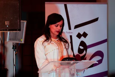 A woman in a white embroidered dress speaks into a microphone at a clear lectern, in front of a banner reading the partial text "My Voice." and Arabic calligraphy.