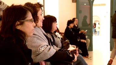 A row of seated people smiling and listening during an indoor gathering, with an open doorway visible in the background.