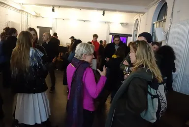 A group of people stand and talk in an indoor space with a lit ceiling, several holding drinks.