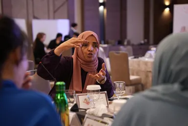 A woman gestures with both hands while speaking at a table set with a green bottle, water glasses and a name card showing a butterfly, with two people in head coverings blurred in the foreground.