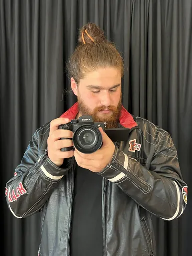 A bearded man with his hair tied up holds a camera with a large lens in front of dark curtains.