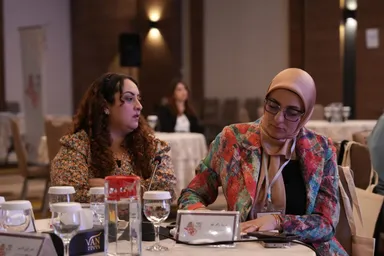 Two women sit at a table with water glasses and a name card reading "VAN", one wearing a colorful patterned jacket and looking down at a phone.