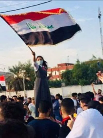 A woman stands above a crowd on someone's shoulders, waving a large flag with a national emblem, surrounded by people raising their arms.