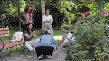 A group of people gathered around a rose bush in a garden, one taking a photo with a phone.