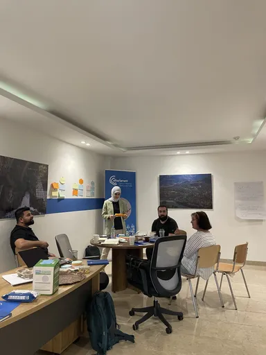 A woman presents notes to three colleagues seated around a table in an office with framed nature photographs and a banner reading "elbarlament cultures of democracy".
