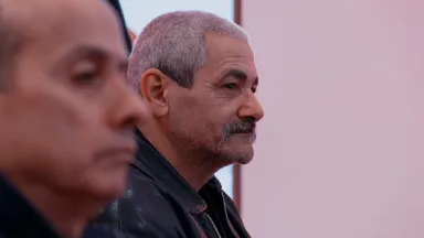 Two men, one in the foreground out of focus, looking off to the side against a plain pink-toned wall.