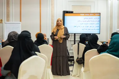 A woman in a brown dress and headscarf addresses a seated group of women in face-covering veils, standing before a screen with Arabic text and a whiteboard.