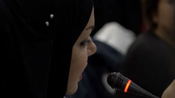 Close-up profile of a person in a headscarf next to a microphone with a lit indicator ring, in a dim room.