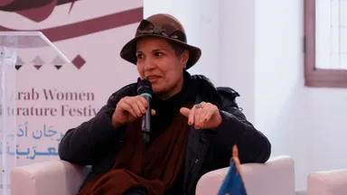 A woman wearing a hat speaking into a microphone and gesturing with her hand, beside a banner reading "Arab Women Literature Festival".
