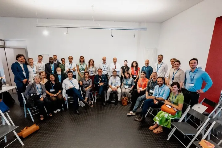 A group of people pose for a photo indoors, some seated and some standing, wearing lanyards, with one face blurred.