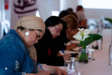 A woman wearing a headscarf writes at a long table set with a flower vase, water bottles and glasses, others seated nearby.