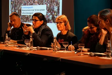 Five people sit at a panel discussion table with microphones and water glasses, in front of a screen reading "8. Werkstattgespräch: Massenproteste im Irak: Teil einer globalen Aufbruchsbewegung?".