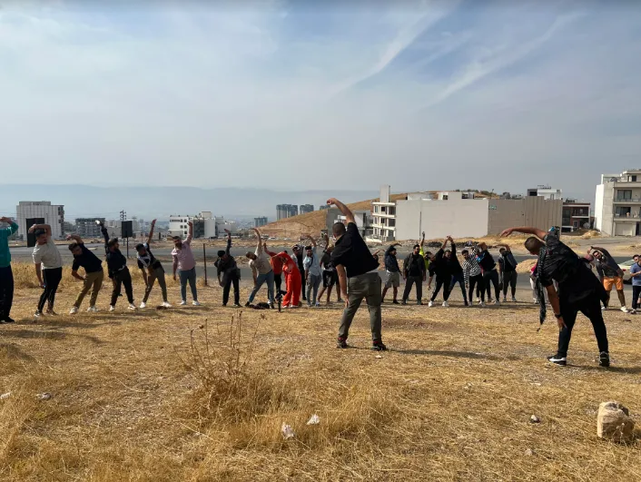 A group of people doing a stretching exercise outdoors on a hillside with a city skyline in the background.