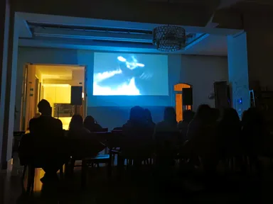 A group of people sit watching video footage projected on a screen in a dimly lit room.