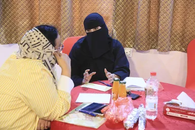 Two women sit at a round table with drinks and papers, one wearing a face-covering veil and gesturing while speaking to the other.