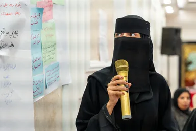A woman in a face-covering veil holds a gold microphone in front of a wall covered with colored sticky notes bearing handwritten Arabic text.