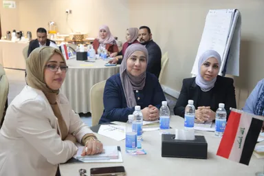 A group of people sit around conference tables with water bottles and an Iraqi flag, with a flip chart in the background.