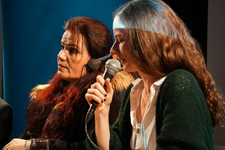 Two women sit close together at a panel discussion, one speaking into a microphone.