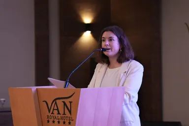 A woman speaks into a microphone at a wooden lectern reading "VAN ROYAL HOTEL" with five stars, with wall lights in the background.