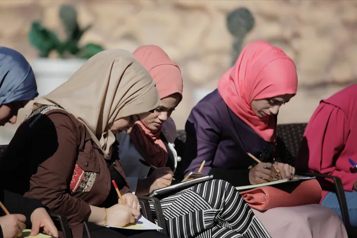 Several women wearing headscarves sit close together, writing on notepads.