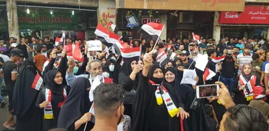 Large crowd waving Iraqi flags at a street protest, with several people holding white signs, in front of shopfronts including one reading "عالم الرياض".