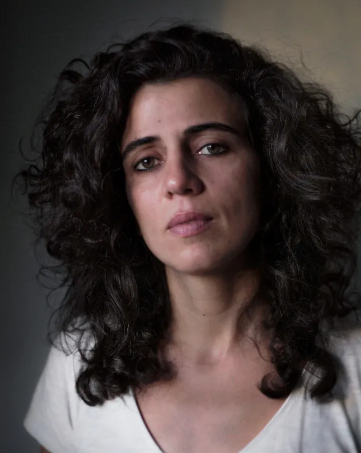 Close-up portrait of a woman with curly dark hair wearing a white top against a plain dark background.