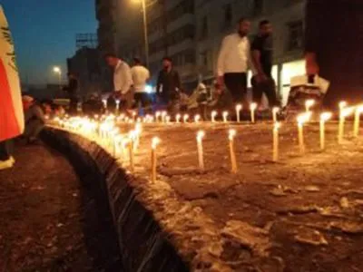 Rows of lit candles line a street edge at night as people walk past in the dim light.