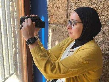 A woman in a headscarf holds a small video camera up to her eye near a window with metal bars.