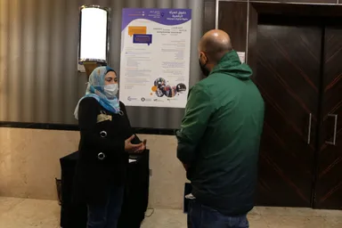 A woman wearing a face mask talks with a man beside a poster reading "Women's Digital Rights" and "حقوق المرأة الرقمية".