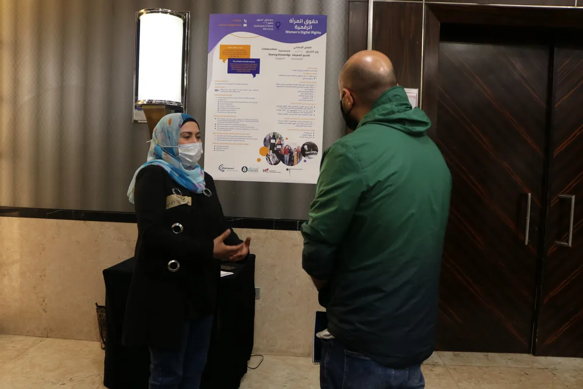 A woman wearing a face mask talks with a man beside a poster reading "Women's Digital Rights" and "حقوق المرأة الرقمية".