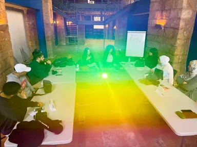 A group of people sit at tables in a vaulted stone hall, with a green light beam shining down and a flip chart in the background.