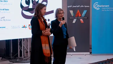 Two women stand together, one speaking into a microphone, in front of banners reading "WORLD ART CONNECTION" and "elbarlament cultures of democracy".