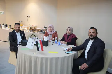 Two men and two women sit at a round table with an Iraqi flag and a flag reading "CORAL".