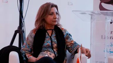 A woman sitting in an armchair wearing a floral top and black scarf, seated beside a lectern with a partially visible event poster.