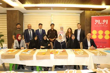 A group of people posing around tables set with tote bags in a conference room, beside a banner reading "OLIRAQ" and "CIVIC SPACE FOR KNOWLEDGE".