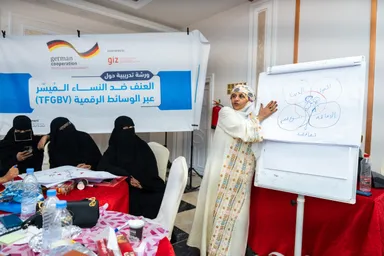 A woman in a headscarf points to a whiteboard with a diagram of connected circles, beside a banner reading "العنف ضد النساء الميسّر عبر الوسائط الرقمية (TFGBV)", with women seated at a table in the foreground.