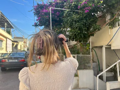 A woman photographs purple flowering branches with a camera, seen from behind on a residential street.
