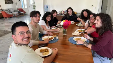 A group of people sit around a wooden table sharing a meal, with a bouquet of colorful flowers in the center.