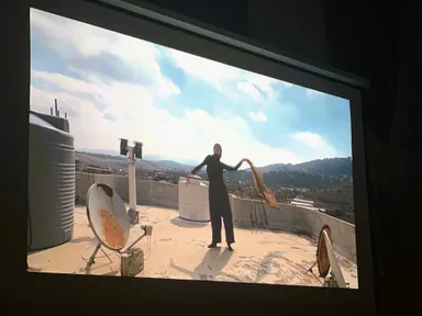 Projected video image of a person in black clothing standing on a rooftop holding a fabric scarf, surrounded by satellite dishes and water tanks with hills in the background.