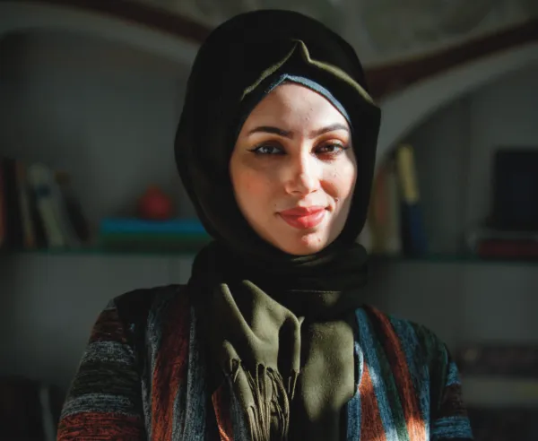 A woman in a black headscarf with a green shawl stands in front of bookshelves.