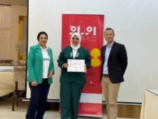 Two women and a man pose indoors in front of a red banner partly reading "...RAQ", one woman holding a certificate.