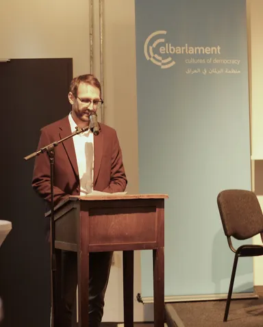 A man reads from papers at a wooden lectern with a microphone, beside a banner reading "elbarlament cultures of democracy" and Arabic text, with an empty chair nearby.