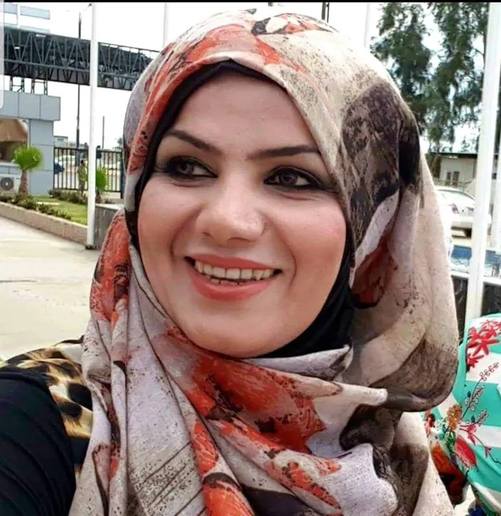 Close-up of a woman wearing a floral headscarf and smiling outdoors, with a building, trees and a car visible behind her.