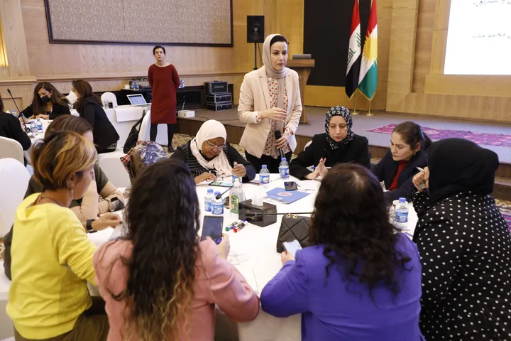 Women seated around a table in discussion, with two women standing at the front and flags and a projection screen behind them.
