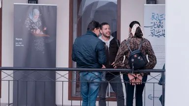 Three people talking outdoors on a terrace, beside exhibition banners showing a portrait photograph with a biographical caption and Arabic calligraphy, next to an "elbarlament" logo.