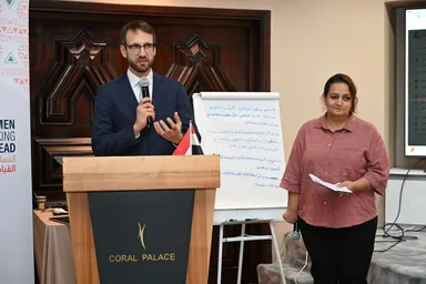A man speaking into a microphone at a podium reading "CORAL PALACE", beside a woman holding papers, with a flip chart of handwritten notes in the background.
