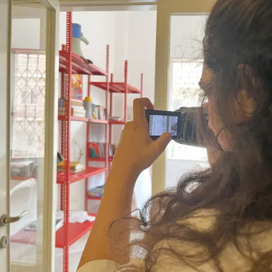 A person photographs a red shelving unit through a doorway using a small camera.