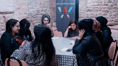 A group of women sitting around a table in a stone-walled room, next to a banner reading "World Art Connection" with Arabic text above it.