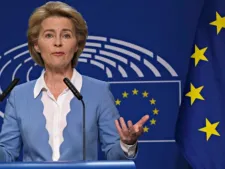 A woman speaks at a podium gesturing with one hand, in front of a European Parliament backdrop and European Union flag.